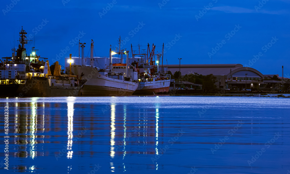 Large oil tanker and fishing boats dock at harbor in industrial area along the river at night