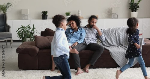 Tired helpless African ethnicity couple sit on sofa feel exhaustion while noisy hyperactive little children running around. Upbringing difficulties, misbehaved son and daughter, ADHD problem concept