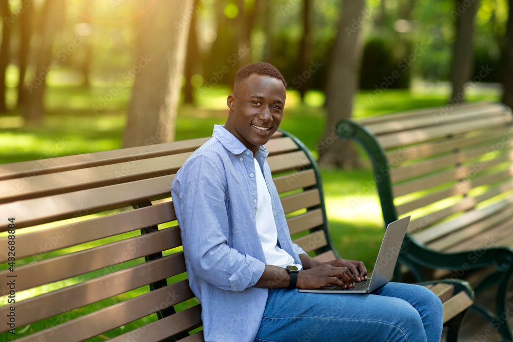 Happy black guy with laptop computer working or studying online on bench at park Happy black guy with laptop computer working or studying online on bench at park