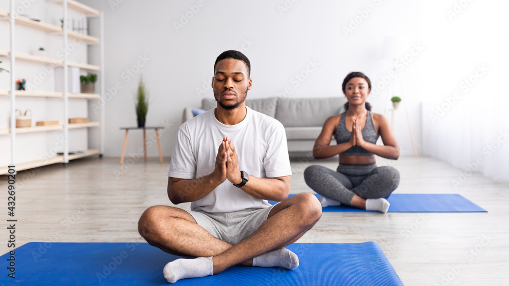 Black guy meditating with closed eyes, sitting in lotus pose, doing namaste gesture, practicing yoga with his girlfriend - obrazy, fototapety, plakaty 