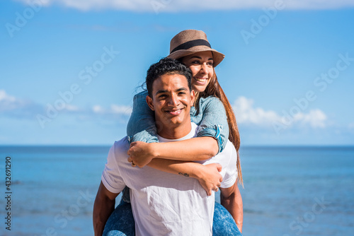 Smiling young man carrying woman on his back and laughing outdoors - young couple people in piggyback have fun together with blue ocean and sky in background