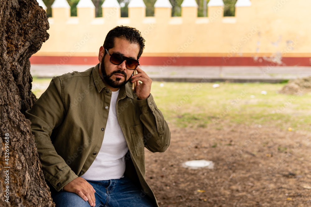 Young bearded serious mexican man wearing jeans and jacket talking on his smartphone and sitting on a tree inside a convent with yellow walls in Mexico. Young bearded serious mexican man wearing jeans and jacket talking on his smartphone and sitting on a tree inside a convent with yellow walls in Mexico.