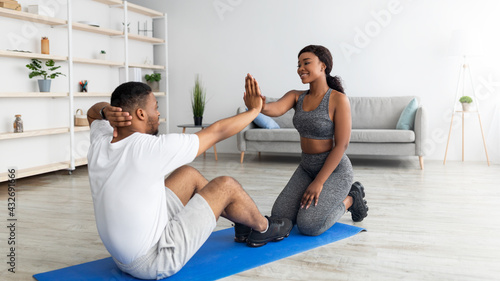 Young black guy doing abs exercises with his girlfriend, high fiving her, working out as team at home, copy space