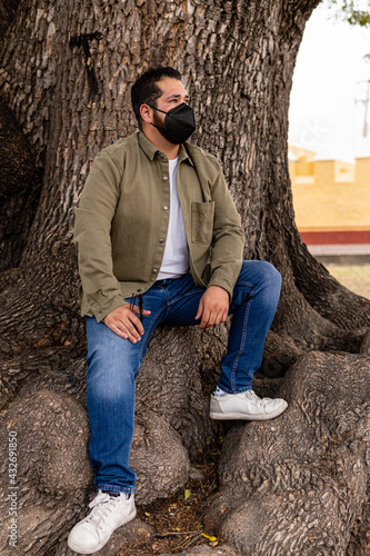 Young overweight mexican man wearing a face mask and a green jacket sitting in a very large tree trunk. Vertical full body portrait