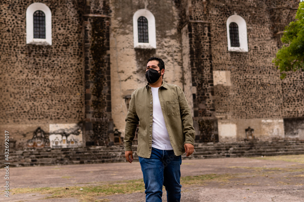 Young bearded mexican man wearing green jacket and face mask walking in front of a church in Mexico. Full Body Portrait