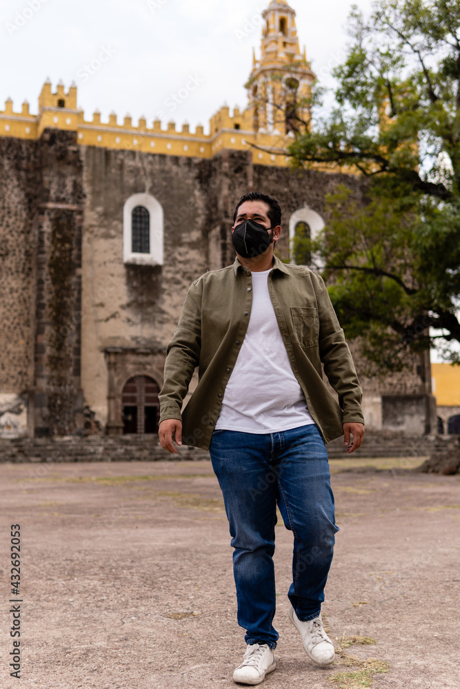 Young bearded mexican man wearing green jacket and face mask walking in front of a church in Mexico. Full Body Portrait