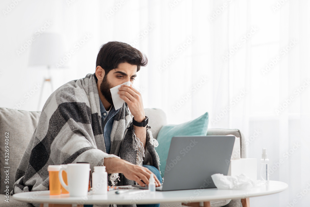 Ill arab guy sneezing and blowing runny nose in paper tissue, working distantly on laptop computer, sitting on sofa Ill arab guy sneezing and blowing runny nose in paper tissue, working distantly on laptop computer, sitting on sofa