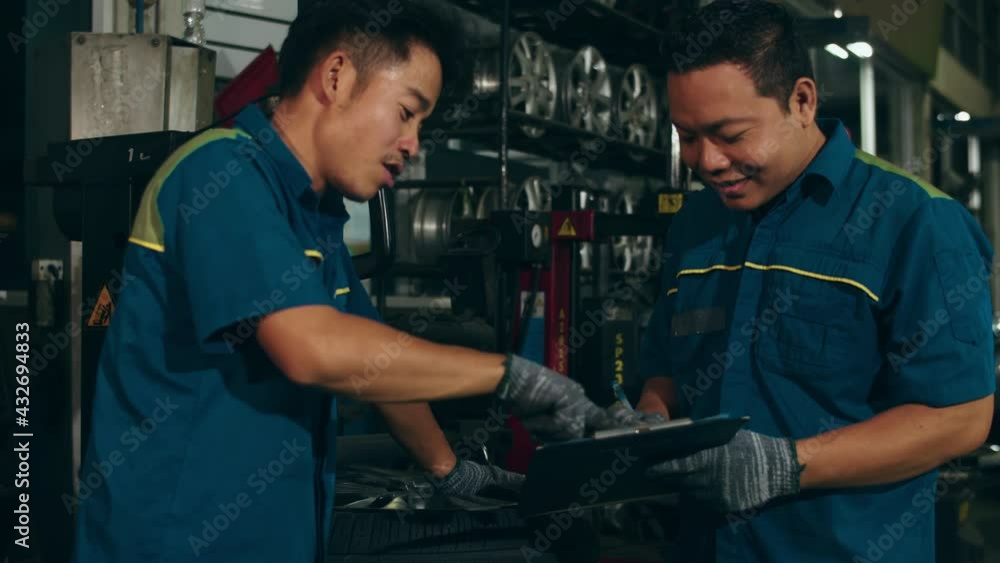 Two professional car mechanic using paperwork makes the oil and engine check to the car on lifted automobile at repair service station at night. Skillful Asian guy in uniform fixing car. Car service. Two professional car mechanic using paperwork makes the oil and engine check to the car on lifted automobile at repair service station at night. Skillful Asian guy in uniform fixing car. Car service.