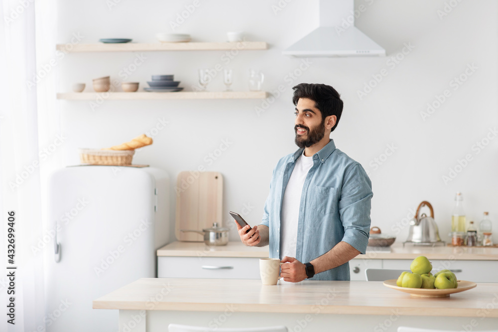 Happy arab guy holding smartphone and drinking coffee, enjoying morning and looking at window in kitchen