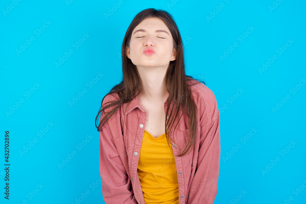 young beautiful Caucasian woman wearing pink jacket over blue wall, keeps lips as going to kiss someone, has glad expression, grimace face. Standing indoors. Beauty concept. young beautiful Caucasian woman wearing pink jacket over blue wall, keeps lips as going to kiss someone, has glad expression, grimace face. Standing indoors. Beauty concept.
