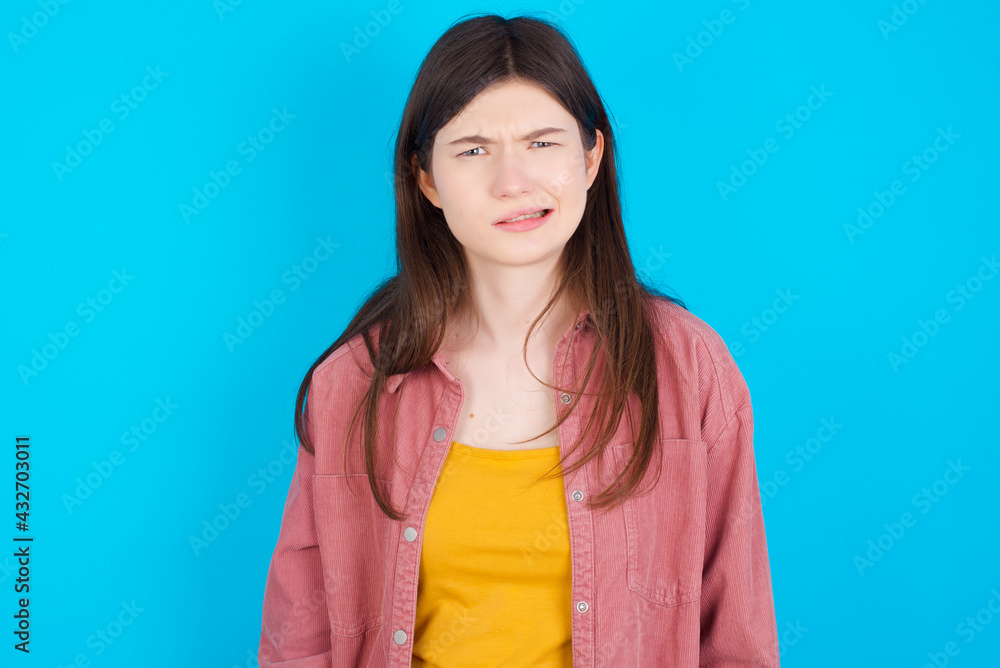young beautiful Caucasian woman wearing pink jacket over blue wall being nervous and scared biting lips looking camera with impatient expression, pensive. young beautiful Caucasian woman wearing pink jacket over blue wall being nervous and scared biting lips looking camera with impatient expression, pensive.