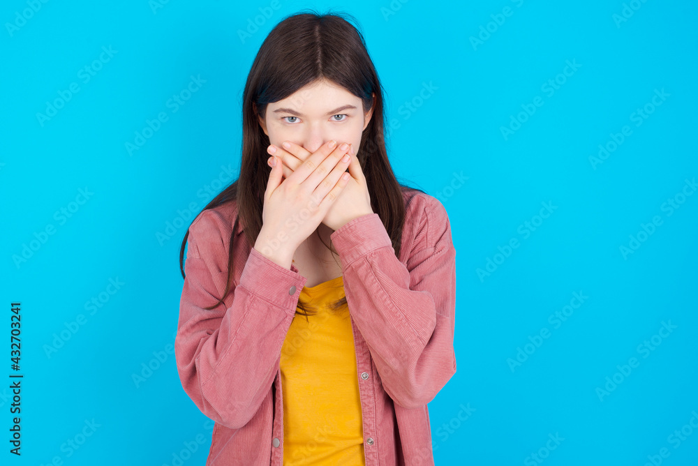 Upset young beautiful Caucasian woman wearing pink jacket over blue wall, covering her mouth with both palms to prevent screaming sound, after seeing or hearing something bad. Upset young beautiful Caucasian woman wearing pink jacket over blue wall, covering her mouth with both palms to prevent screaming sound, after seeing or hearing something bad.