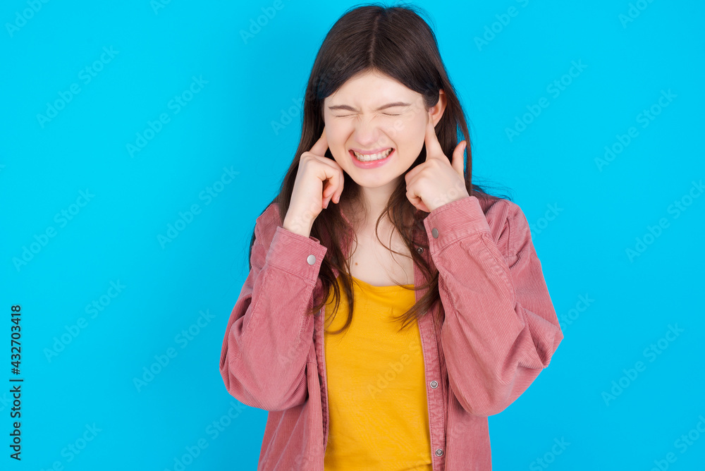 Stop making this annoying sound! Unhappy stressed out young beautiful Caucasian woman wearing pink jacket over blue wall making worry face, plugging ears with fingers, irritated with loud noise. Stop making this annoying sound! Unhappy stressed out young beautiful Caucasian woman wearing pink jacket over blue wall making worry face, plugging ears with fingers, irritated with loud noise.