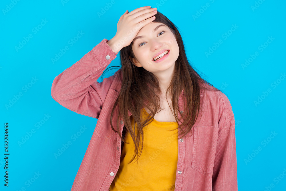 Oops, what did I do? young beautiful Caucasian woman wearing pink T-shirt over blue wall holding hand on forehead with frightened and regret expression. Oops, what did I do? young beautiful Caucasian woman wearing pink T-shirt over blue wall holding hand on forehead with frightened and regret expression.