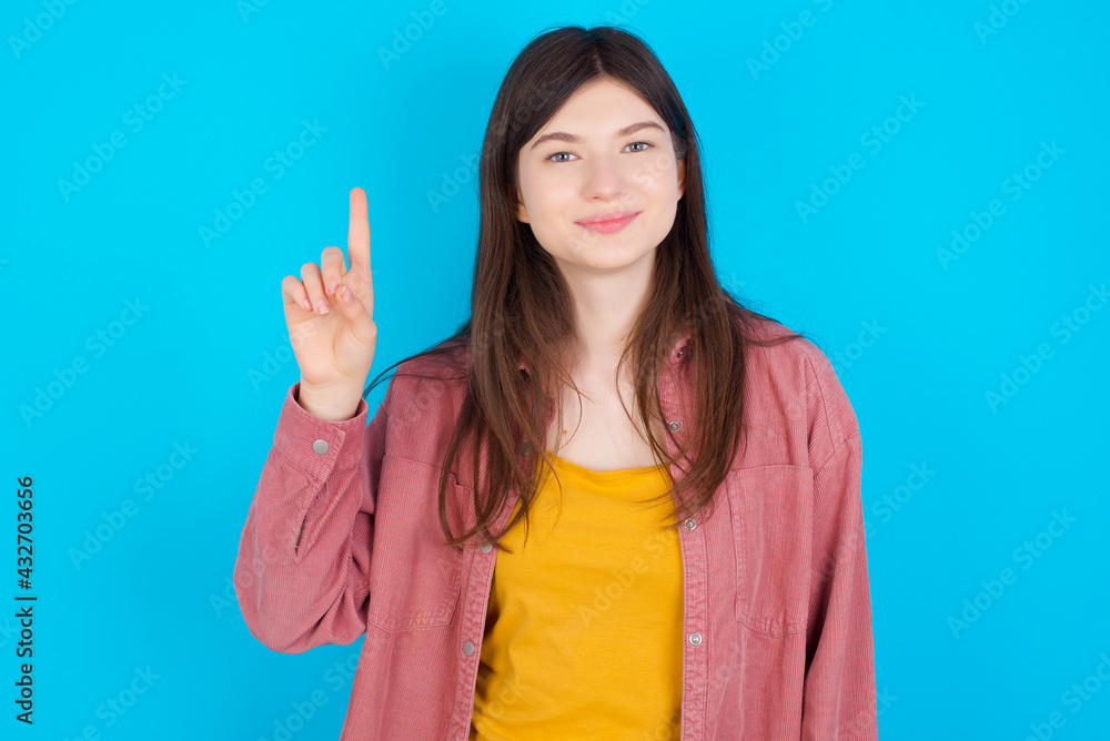 young beautiful Caucasian woman wearing pink T-shirt over blue wall showing and pointing up with fingers number one while smiling confident and happy. young beautiful Caucasian woman wearing pink T-shirt over blue wall showing and pointing up with fingers number one while smiling confident and happy.