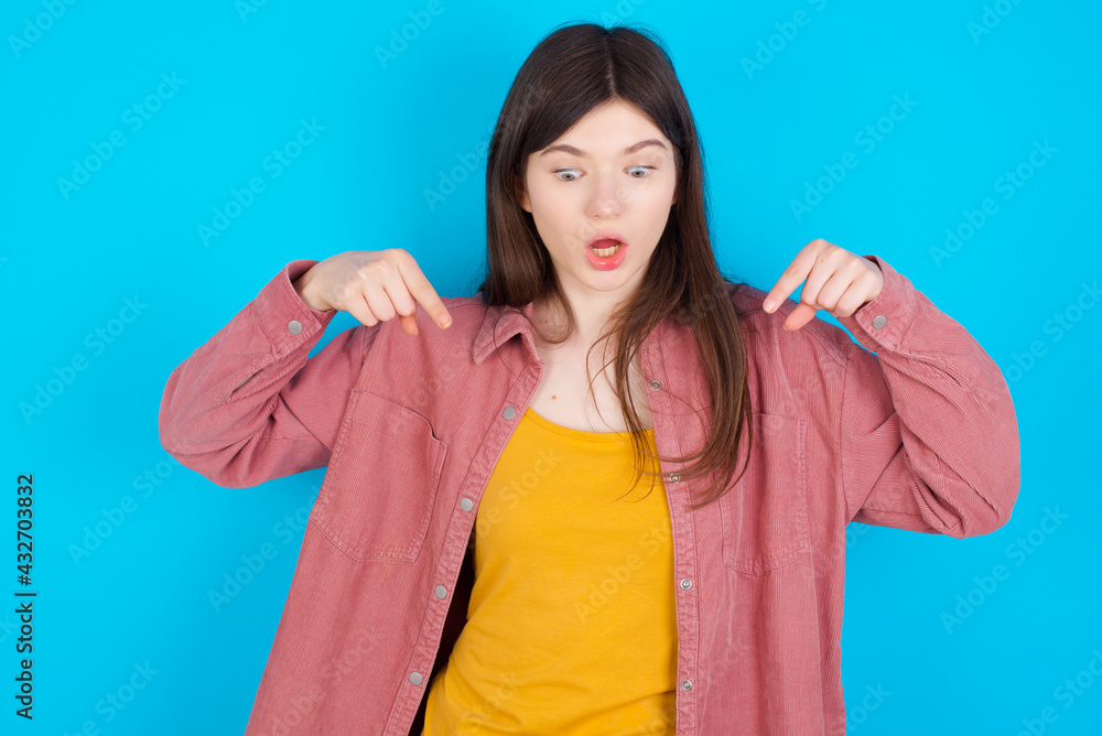 Amazed young beautiful Caucasian woman wearing pink T-shirt over blue wall points down with fore fingers, opens mouth being shocked. Advertisement concept. Amazed young beautiful Caucasian woman wearing pink T-shirt over blue wall points down with fore fingers, opens mouth being shocked. Advertisement concept.