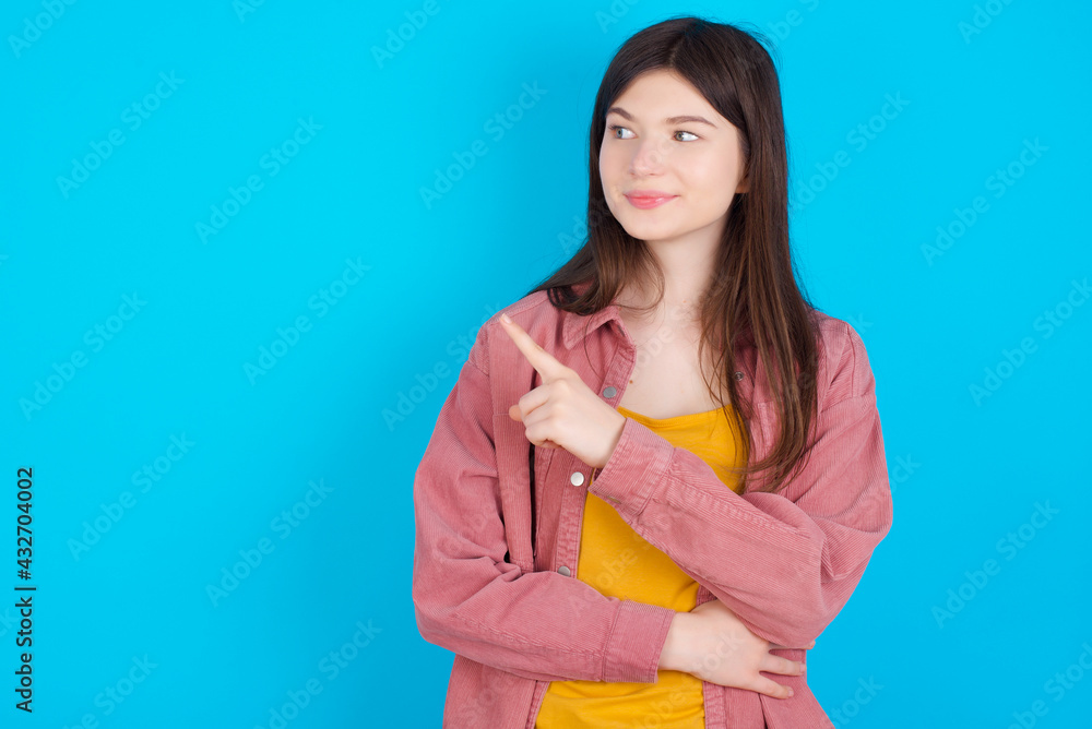 Portrait of young beautiful Caucasian woman wearing pink jacket over blue wall posing on camera with tricky look, presenting product with index finger. Advertisement concept. Portrait of young beautiful Caucasian woman wearing pink jacket over blue wall posing on camera with tricky look, presenting product with index finger. Advertisement concept.