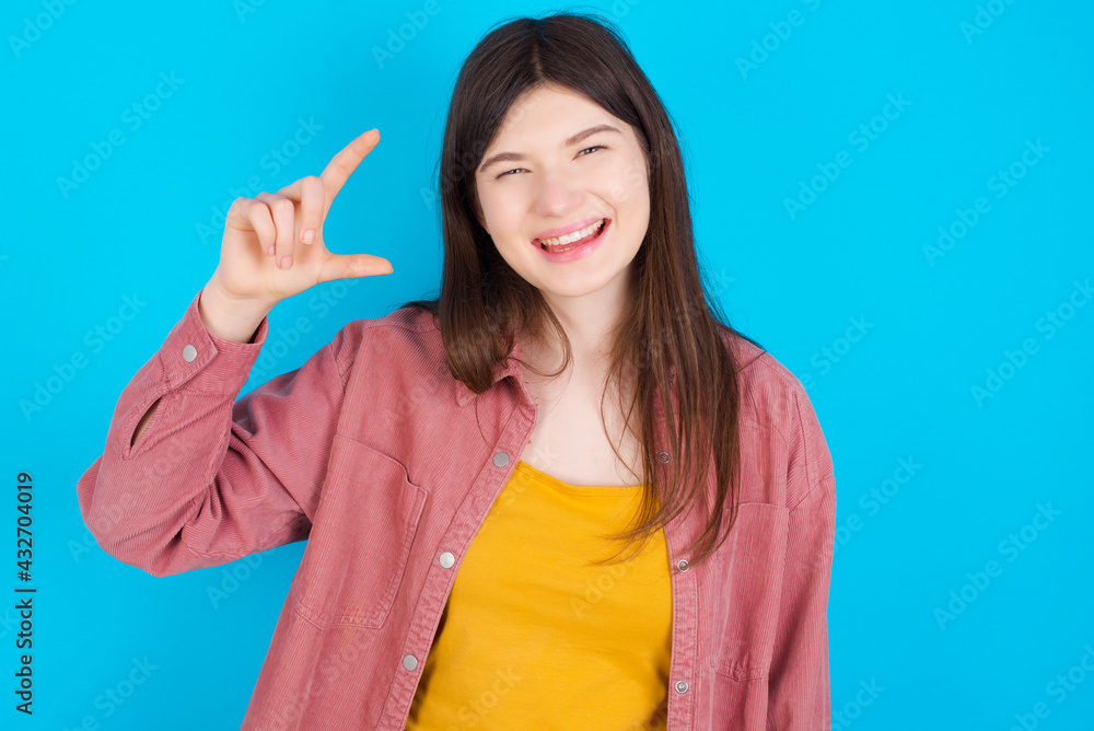 young beautiful Caucasian woman wearing pink jacket over blue wall smiling and gesturing with hand small size, measure symbol. young beautiful Caucasian woman wearing pink jacket over blue wall smiling and gesturing with hand small size, measure symbol.
