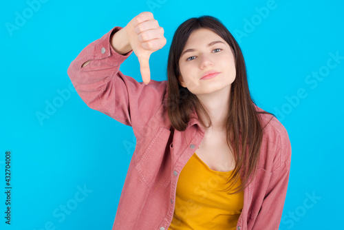 Discontent young beautiful Caucasian woman wearing pink jacket over blue wall shows disapproval sign, keeps thumb down, expresses dislike, frowns face in discontent. Negative feelings.
