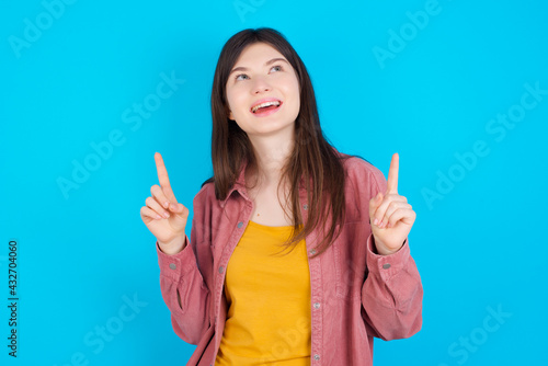 Successful friendly looking young beautiful Caucasian woman wearing pink jacket over blue wall exclaiming excitedly, pointing both index fingers up, indicating something.