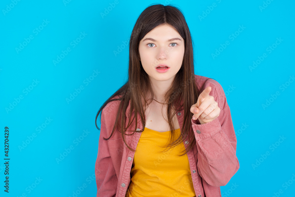 Shocked young beautiful Caucasian woman wearing pink jacket over blue wall points front with index finger at camera and. Surprise and advertisement concept. Shocked young beautiful Caucasian woman wearing pink jacket over blue wall points front with index finger at camera and. Surprise and advertisement concept.