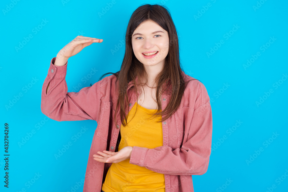 young beautiful Caucasian woman wearing pink jacket over blue wall gesturing with hands showing big and large size sign, measure symbol. young beautiful Caucasian woman wearing pink jacket over blue wall gesturing with hands showing big and large size sign, measure symbol.