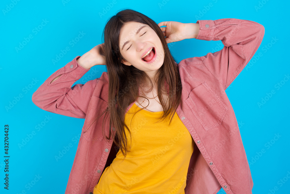 young beautiful Caucasian woman wearing pink jacket over blue wall relaxing and stretching, arms and hands behind head and neck smiling happy young beautiful Caucasian woman wearing pink jacket over blue wall relaxing and stretching, arms and hands behind head and neck smiling happy