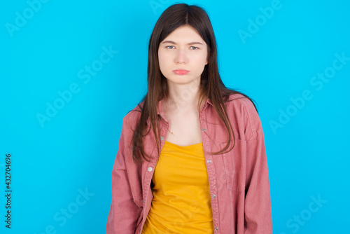 Gloomy, bored young beautiful Caucasian woman wearing pink jacket over blue wall frowns face looking up, being upset with so much talking hands down, feels tired and wants to leave.