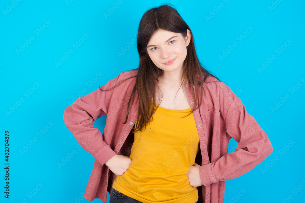 Funny frustrated young beautiful Caucasian woman wearing pink jacket over blue wall holding hands on waist and silly looking at awkward situation. Funny frustrated young beautiful Caucasian woman wearing pink jacket over blue wall holding hands on waist and silly looking at awkward situation.