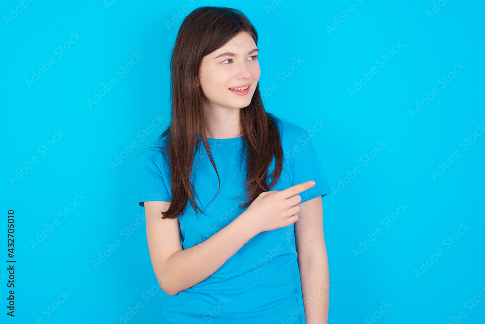 Happy cheerful smiling young beautiful Caucasian woman wearing blue T-shirt over blue wall looking and pointing aside with hand. Copy space and advertisement concept. Happy cheerful smiling young beautiful Caucasian woman wearing blue T-shirt over blue wall looking and pointing aside with hand. Copy space and advertisement concept.