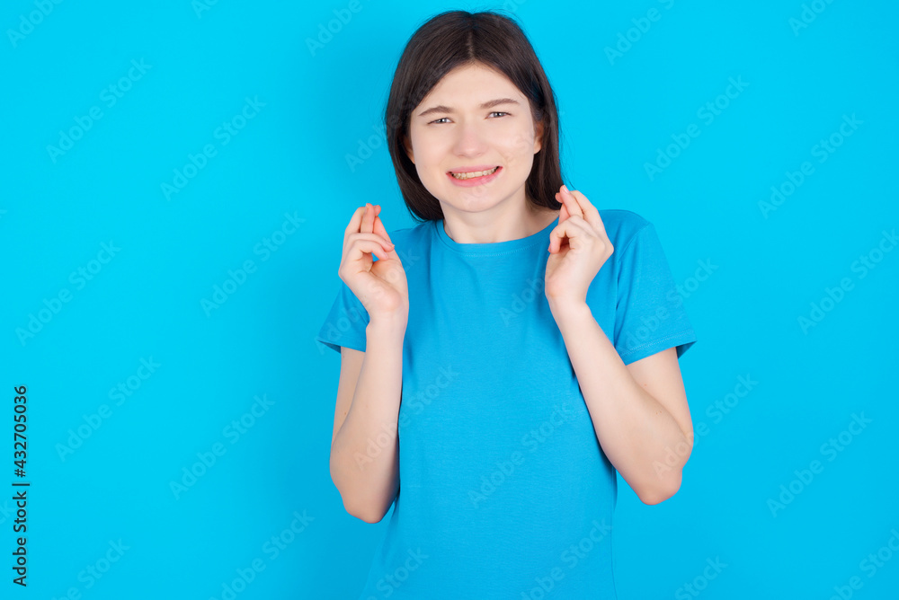 young beautiful Caucasian woman wearing blue T-shirt over blue wall holding fingers crossed with worried expression hoping boss didn't noticed mistakes at work. young beautiful Caucasian woman wearing blue T-shirt over blue wall holding fingers crossed with worried expression hoping boss didn't noticed mistakes at work.