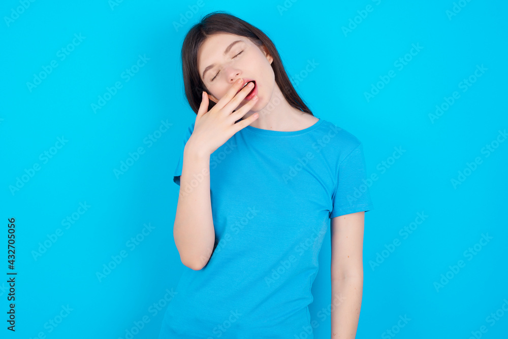 Sleepy young beautiful Caucasian woman wearing blue T-shirt over blue wall yawning with messy hair, feeling tired after sleepless night, yawning, covering mouth with palm. Sleepy young beautiful Caucasian woman wearing blue T-shirt over blue wall yawning with messy hair, feeling tired after sleepless night, yawning, covering mouth with palm.