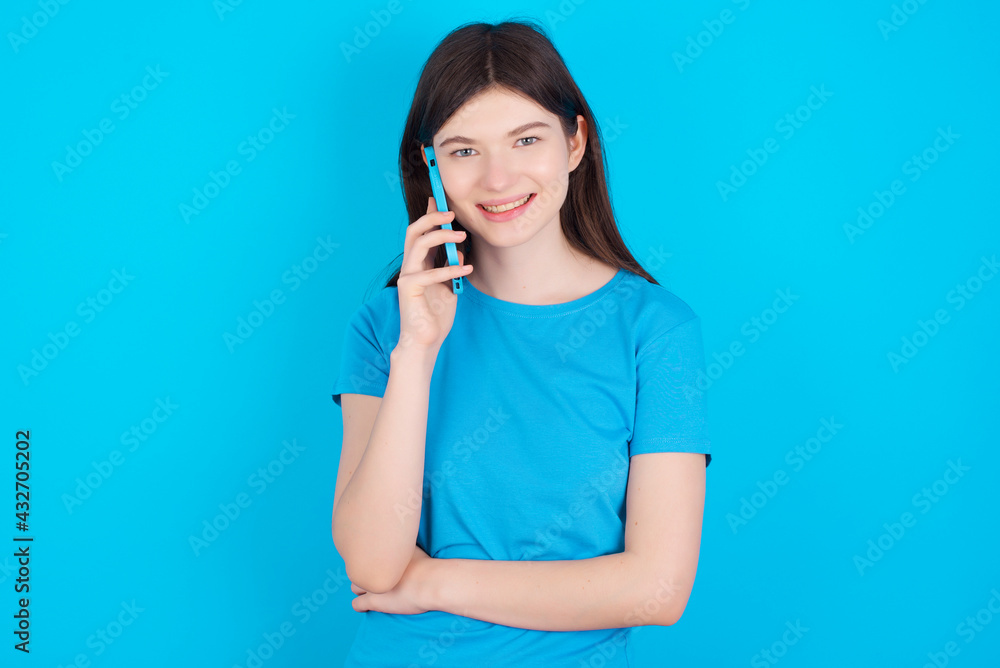Portrait of a smiling young beautiful Caucasian woman wearing blue T-shirt over blue wall talking on mobile phone. Business, confidence and communication concept. Portrait of a smiling young beautiful Caucasian woman wearing blue T-shirt over blue wall talking on mobile phone. Business, confidence and communication concept.