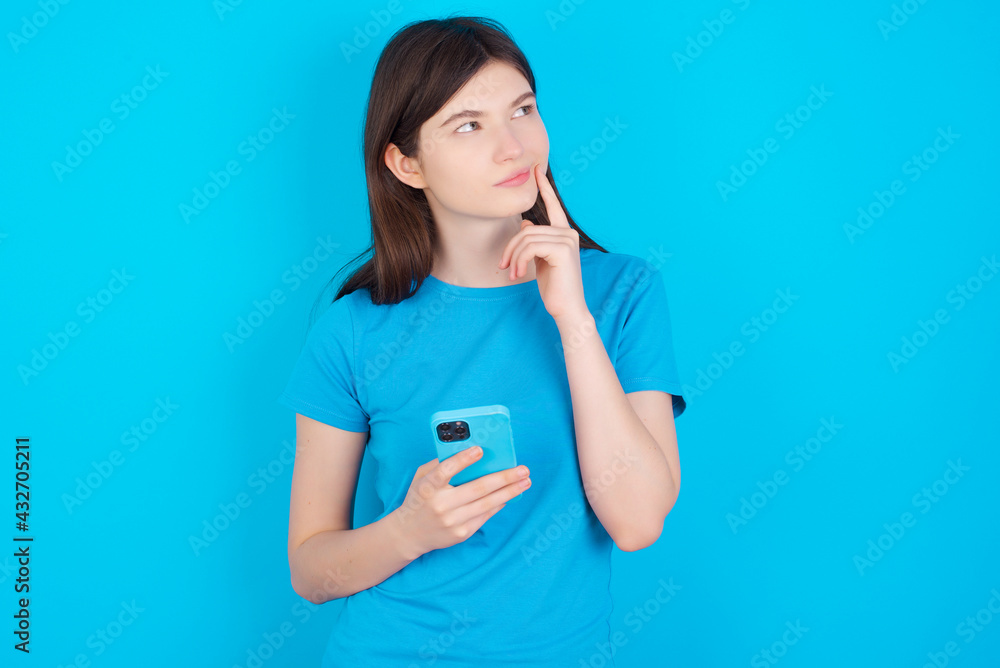 Thinking dreaming young beautiful Caucasian woman wearing blue T-shirt over blue wall using mobile phone and holding hand on face. Taking decisions and social media concept. Thinking dreaming young beautiful Caucasian woman wearing blue T-shirt over blue wall using mobile phone and holding hand on face. Taking decisions and social media concept.