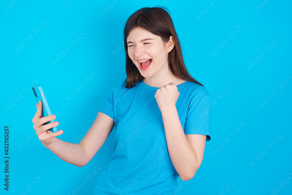 Happy cheerful young beautiful Caucasian woman wearing blue T-shirt over blue wall receiving good news via e-mail and celebrating success while standing and looking at mobile phone.