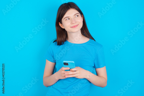 Happy young beautiful Caucasian woman wearing blue T-shirt over blue wall listening to music with earphones using mobile phone.