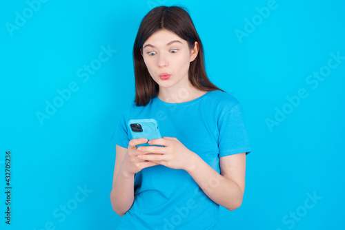 young beautiful Caucasian woman wearing blue T-shirt over blue wall looks with bugged eyes, holds modern smart phone, receives unexpected message from friend, reads reminder.