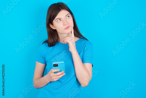 Image of a thinking dreaming young beautiful Caucasian woman wearing blue T-shirt over blue wall using mobile phone and holding hand on face. Taking decisions and social media concept.