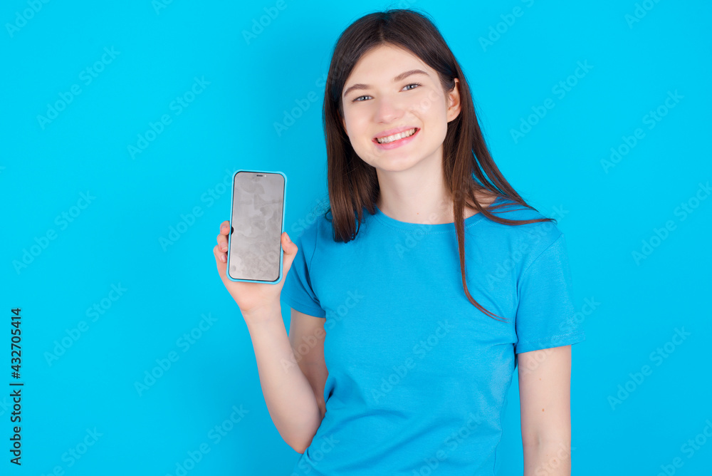 Smiling young beautiful Caucasian woman wearing blue T-shirt over blue wall showing empty phone screen. Advertisement and communication concept. Smiling young beautiful Caucasian woman wearing blue T-shirt over blue wall showing empty phone screen. Advertisement and communication concept.