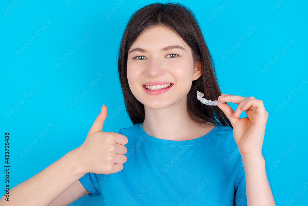 young beautiful Caucasian woman wearing blue T-shirt over blue wall holding an invisible braces aligner and rising thumb up, recommending this new treatment. Dental healthcare concept.