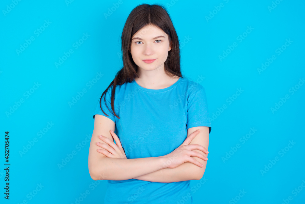 Self confident serious calm young beautiful Caucasian woman wearing blue T-shirt over blue wall stands with arms folded. Shows professional vibe stands in assertive pose. Self confident serious calm young beautiful Caucasian woman wearing blue T-shirt over blue wall stands with arms folded. Shows professional vibe stands in assertive pose.