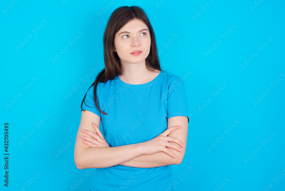 Charming thoughtful young beautiful Caucasian woman wearing blue T-shirt over blue wall stands with arms folded concentrated somewhere with pensive expression thinks what to do