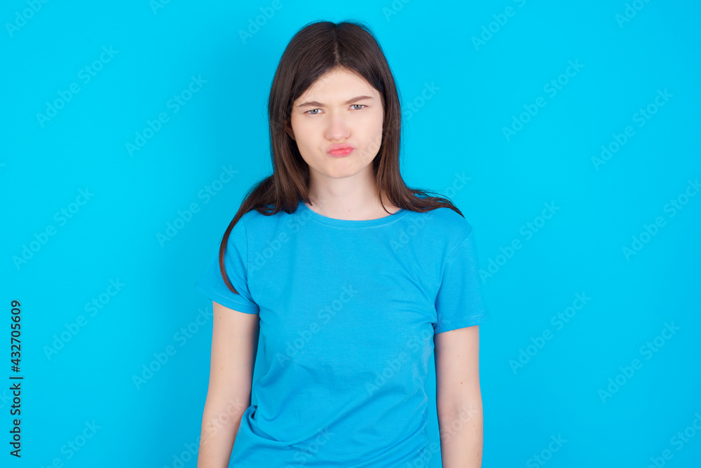 Offended dissatisfied young beautiful Caucasian woman wearing blue T-shirt over blue wall with moody displeased expression at camera being disappointed by something Offended dissatisfied young beautiful Caucasian woman wearing blue T-shirt over blue wall with moody displeased expression at camera being disappointed by something