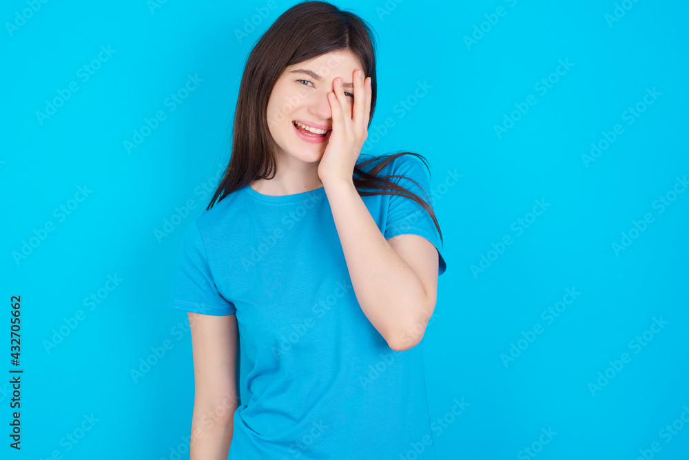 young beautiful Caucasian woman wearing blue T-shirt over blue wall makes face palm and smiles broadly, giggles positively hears funny joke poses young beautiful Caucasian woman wearing blue T-shirt over blue wall makes face palm and smiles broadly, giggles positively hears funny joke poses