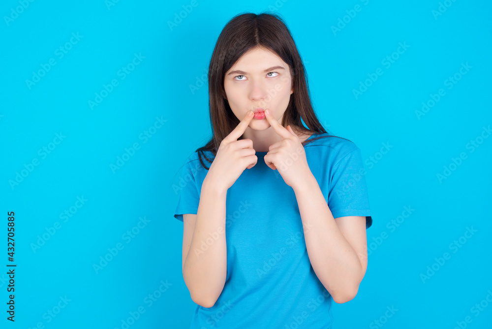 young beautiful Caucasian woman wearing blue T-shirt over blue wall crosses eyes and makes fish lips funny grimace young beautiful Caucasian woman wearing blue T-shirt over blue wall crosses eyes and makes fish lips funny grimace