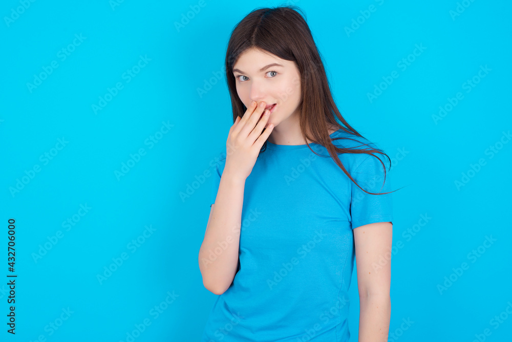 young beautiful Caucasian woman wearing blue T-shirt over blue wall covers mouth and looks with wonder at camera, cannot believe unexpected rumors. young beautiful Caucasian woman wearing blue T-shirt over blue wall covers mouth and looks with wonder at camera, cannot believe unexpected rumors.