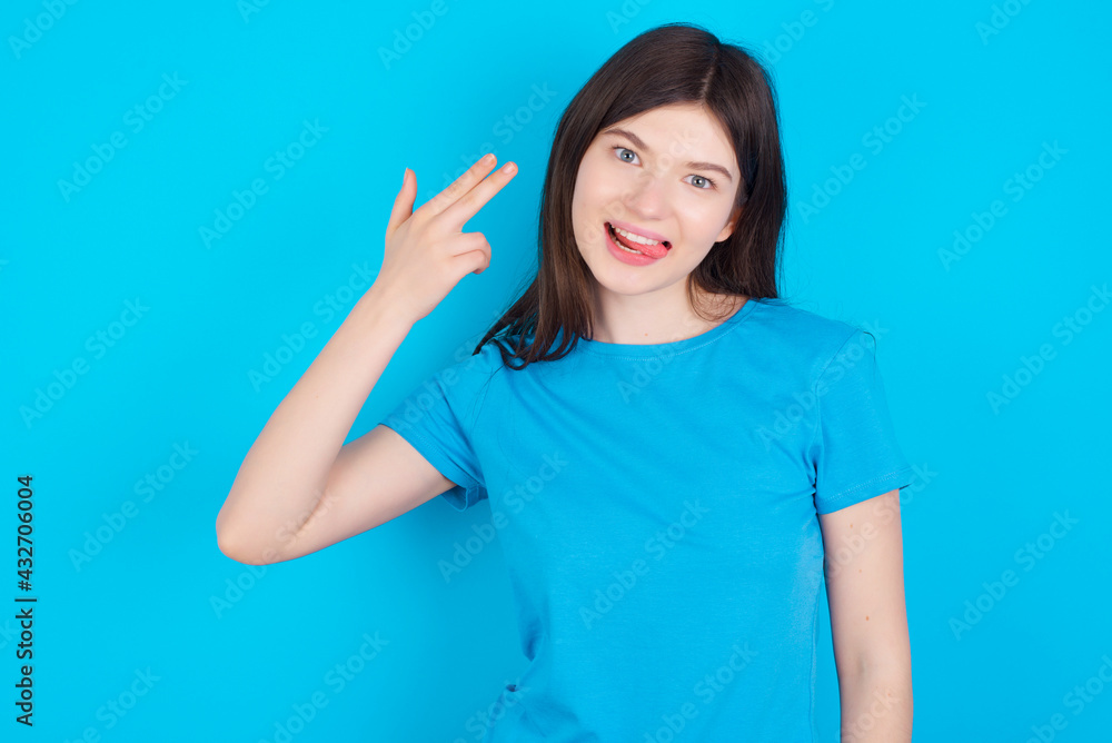 young beautiful Caucasian woman wearing blue T-shirt over blue wall foolishes around shoots in temple with fingers makes suicide gesture. Funny model makes finger gun pistol young beautiful Caucasian woman wearing blue T-shirt over blue wall foolishes around shoots in temple with fingers makes suicide gesture. Funny model makes finger gun pistol