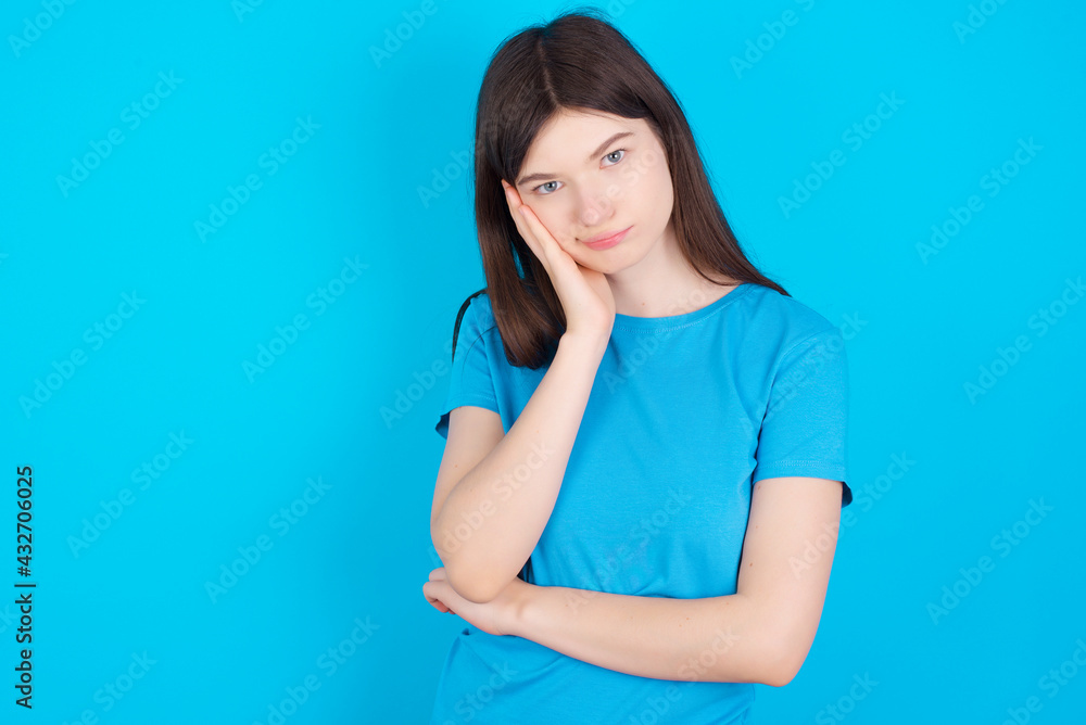 Sad lonely young beautiful Caucasian woman wearing blue T-shirt over blue wall touches cheek with hand bites lower lip and gazes with displeasure. Bad emotions Sad lonely young beautiful Caucasian woman wearing blue T-shirt over blue wall touches cheek with hand bites lower lip and gazes with displeasure. Bad emotions