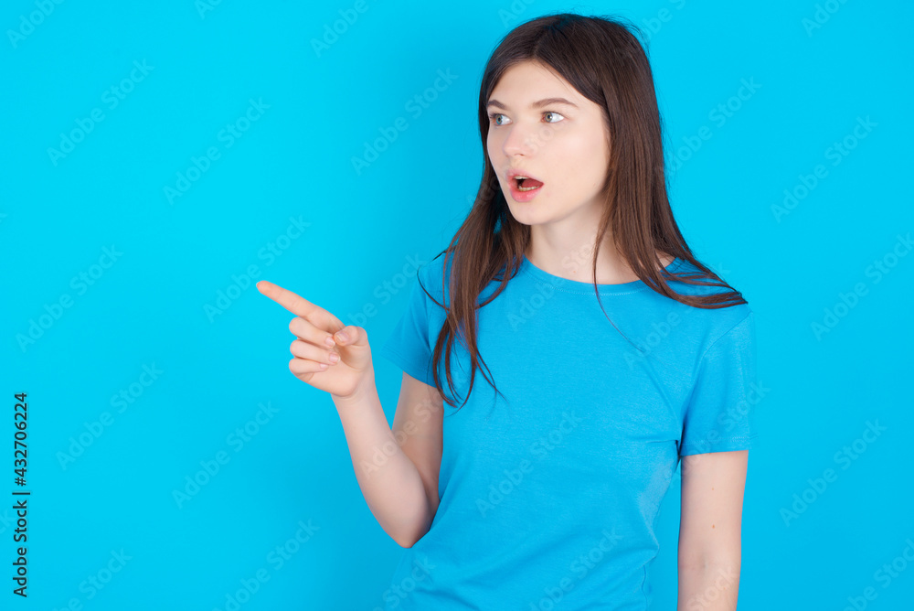 Stunned young beautiful Caucasian woman wearing blue T-shirt over blue wall with greatly surprised expression points away on copy space, indicates something Stunned young beautiful Caucasian woman wearing blue T-shirt over blue wall with greatly surprised expression points away on copy space, indicates something
