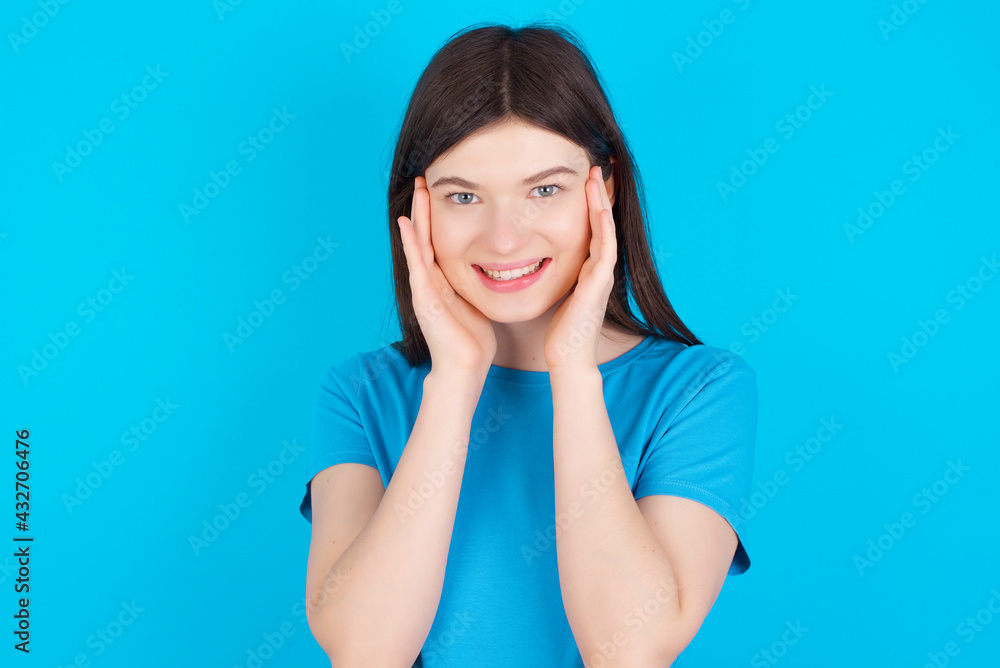 Happy young beautiful Caucasian woman wearing blue T-shirt over blue wall touches both cheeks gently, has tender smile, shows white teeth, gazes positively straightly at camera, Happy young beautiful Caucasian woman wearing blue T-shirt over blue wall touches both cheeks gently, has tender smile, shows white teeth, gazes positively straightly at camera,